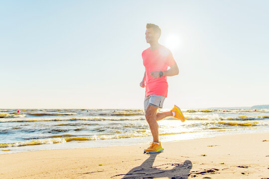 Man jogging along a sandy beach during sunset with waves and a bright sun in the background