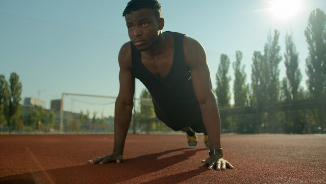 Motivated African American sport man athlete bodybuilder doing push ups exercise on football stadium male young sportsman fitness trainer in city training arms biceps muscles summer workout outdoors