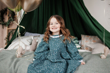 Cheerful little birthday girl 4-5 years in stylish rustic style dress sits on bed decorated with baldachin, pillows and balloons, smiles and looks at camera. Child birthday party celebration. © Andriy Medvediuk