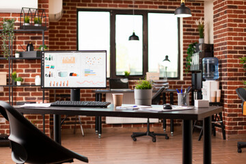 Table in spacious startup office holds desktop pc showing financial analytics. Brick wall interior includes digital devices, shelves, water cooler, and plant decorations, creating refreshing workspace