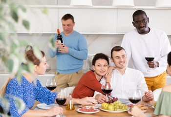 Ethnically diverse adult friends sharing stories and laughing over wine and appetizers in friendly home dinner party setting in cozy kitchen..