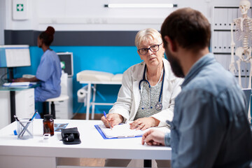 Fototapeta premium Retired female physician with clipboard consulting with young male patient in clinic as nurse works in background. Elderly doctor listening to man with beard and writing his medical symptoms.