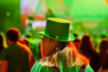 Back view of woman wearing a green hat with glowing stage lights and crowd, St. Patrick's Day...