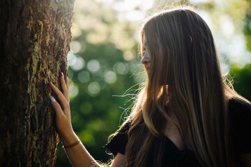 Slender teenage girl caressing tree bark, sunlight filtering through verdant forest foliage
