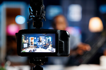 Focus on professional video production camera used for capturing footage of BIPOC content creator in blurry background filming tech reviews for his internet channel, showcasing gaming keyboard