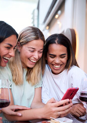 Vertical. Three happy smiling female friends sharing a tablet computer as they stand close together looking at the screen. Multiracial women using mobile smart phone on a terrace bar drinking wine