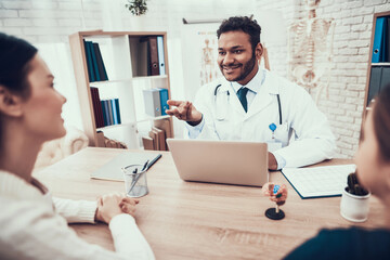 Indian doctor seeing patients in office. Doctor is using laptop at table.