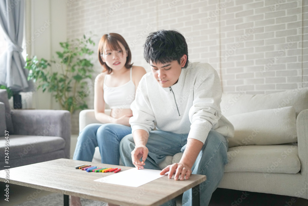 Wall mural A pregnant Japanese woman in her 20s, wearing clothes that reveal her stomach, and her husband are sitting on a sofa in their apartment, drawing a picture.