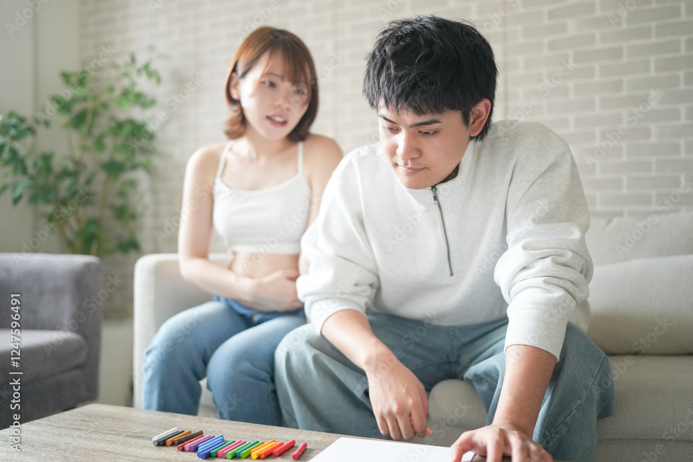 Wall mural A pregnant Japanese woman in her 20s, wearing clothes that reveal her stomach, and her husband are sitting on a sofa in their apartment, drawing a picture.