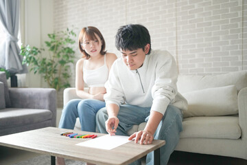 A pregnant Japanese woman in her 20s, wearing clothes that reveal her stomach, and her husband are sitting on a sofa in their apartment, drawing a picture.