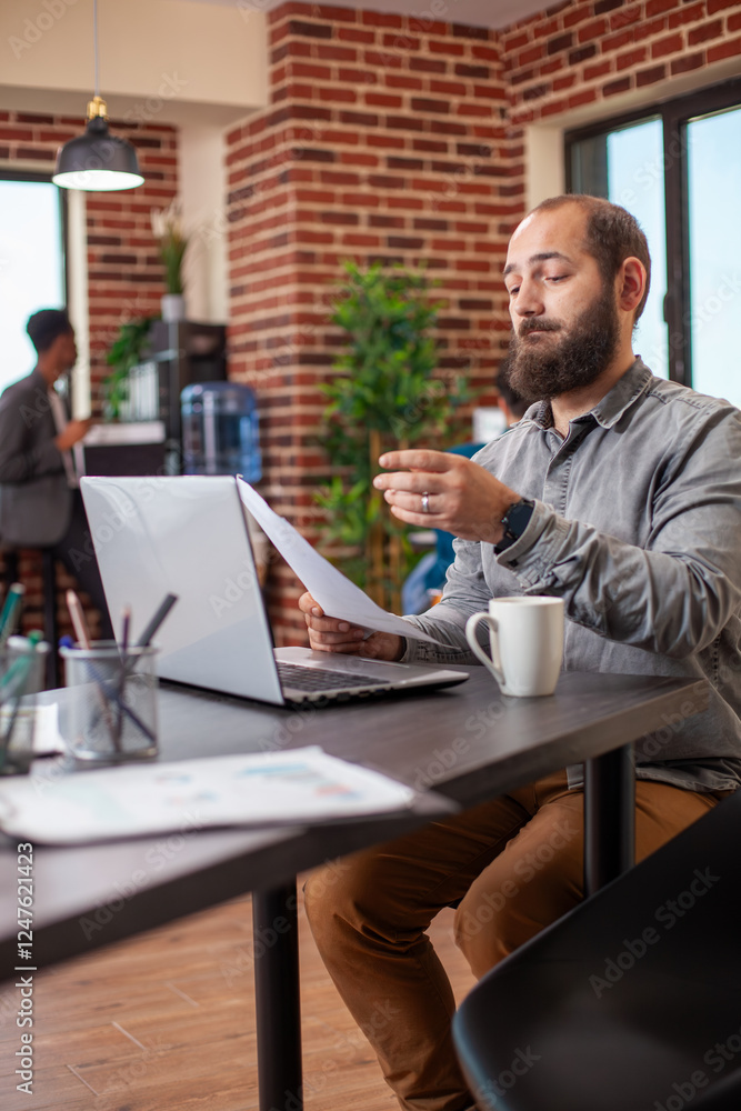 Poster financial specialist seated with laptop is holding a document and reviewing research data in modern 