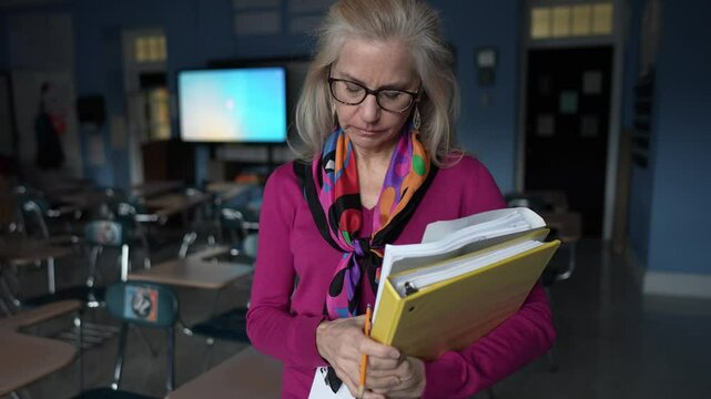 A mature woman stands in an empty classroom holding folders, preparing for her lessons. Her forlorn expression reflects her sadness, frustration in teaching.