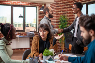 Bearded man shakes hands with African American manager in brick wall office as their colleagues have a discussion at table. Male coworkers stand and exchange greetings during team meeting.