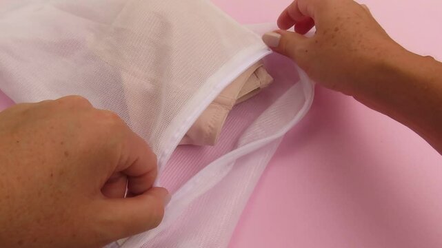 Video of woman hands putting a beige bra into a white laundry bag and closing the zipper on light pink background. Organization concept.