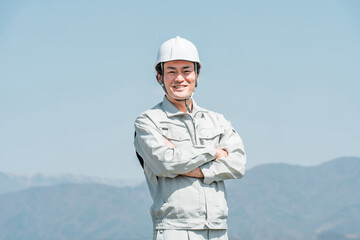 青空の下に立つヘルメットを被った作業着姿の男性（建築・土木・建設）

