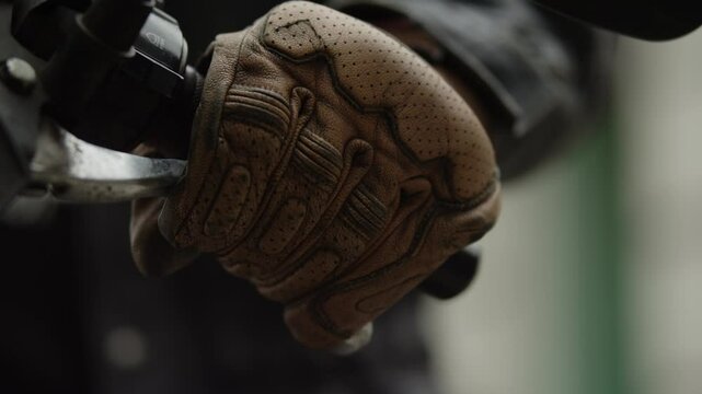 A biker guy sits on a motorcycle during the day, he is wearing brown leather gloves and a black leather jacket, he holds on to the steering wheel, squeezes the clutch lever and slowly releases it