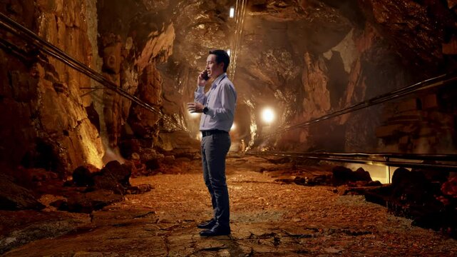 Full Body Side View Of An Asian Male Professional Worker Standing In Underground Mine Tunnel, He Is Speaking With Someone In The Phone And Moving His Left Hand