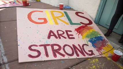 Handmade protest sign with colorful text 'GIRLS ARE STRONG' and rainbow paint splashes on pink background. Feminist activism and empowerment concept