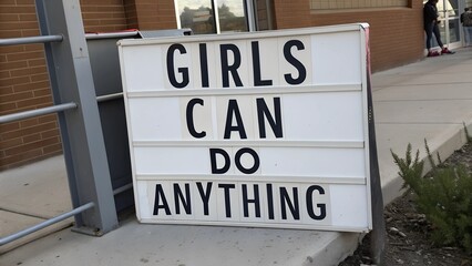 Outdoor white lightbox sign with bold black text 'GIRLS CAN DO ANYTHING' placed on a sidewalk near a brick building. Feminist empowerment and motivation concept