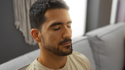 Young hispanic man relaxing indoors with eyes closed, sitting comfortably in a modern home interior, showcasing calm and peaceful living room atmosphere.