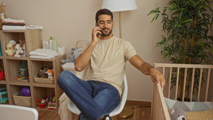 Handsome man with beard sits in bedroom beside baby cradle on phone surrounded by cozy home decor...
