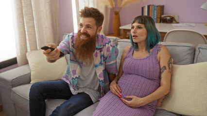 Obraz premium Pregnant woman and man relaxing together in living room at home, smiling and watching television with woman touching her baby belly.