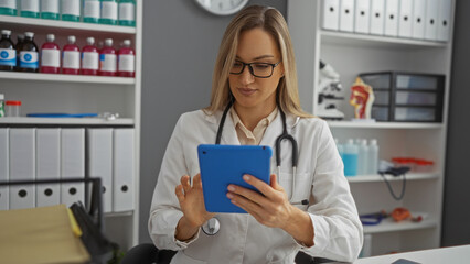 Woman doctor analyzing data on tablet in clinic room with shelves filled with medical supplies and anatomical models, wearing white coat and stethoscope.