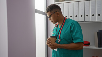 Young hispanic man in medical scrubs stands in a clinic room holding a coffee cup looking thoughtful near a window.