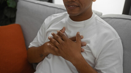 Hand tattooed young hispanic man in white shirt touches chest sitting in cozy apartment living room.