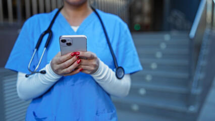 Female nurse in blue scrubs standing outdoors holding smartphone, focusing on screen with stethoscope around neck, city hospital steps in background, daytime.
