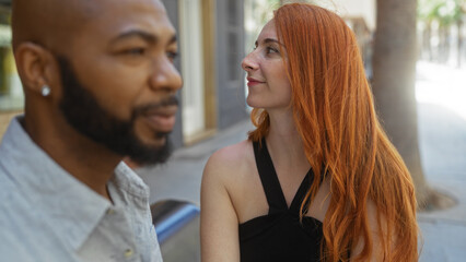 Naklejka premium Interracial couple in love sitting together on an urban street, the woman with red hair smiling at the man