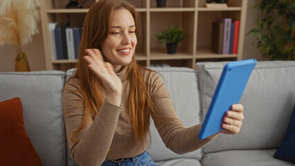 Woman waving on video call using tablet in cozy apartment living room with bookshelf backdrop, showcasing relaxing indoor home environment.