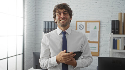 Fototapeta premium Hispanic man in office setting holding tablet with confident smile, exuding professionalism in bright, modern workplace with brick wall and organized background.