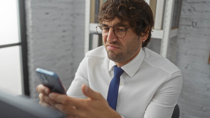 Fototapeta premium Young man frowning at smartphone in bright office setting with glasses, tie, and business attire suggest professional frustration or concern.