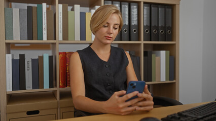 Woman working in office, attractive young blonde female with short hair using smartphone while...