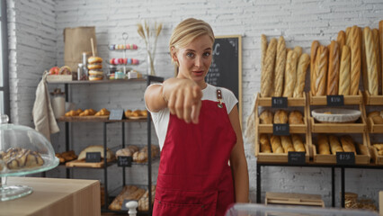 Young, attractive woman with short blonde hair and a red apron pointing at the camera in an indoor bakery filled with various bread and pastries on shelves.