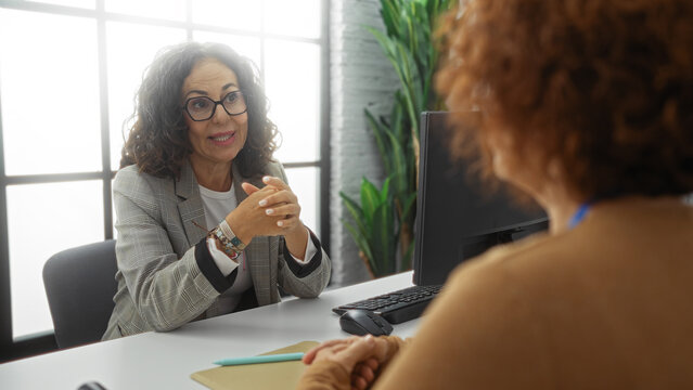 Women discussing business indoors, featuring two mature hispanic workers engaged in professional conversation in a modern office setting.