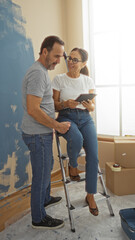 Hispanic couple stands together in a new home living room, painting walls and looking at a tablet,...