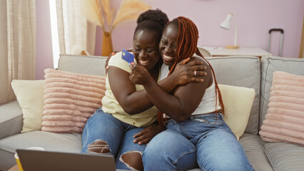 Two happy african american women, sisters or friends, hugging and smiling on a couch in a cozy living room, celebrating with a credit card, indicating love and family bonding.