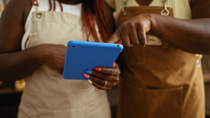 Two black women bakers in a bakery shop are using a blue tablet together, focusing on work and collaboration in an indoor setting with various equipment in the background.