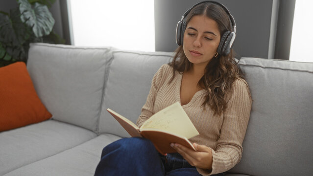Woman relaxing on a sofa with headphones reading a book in an apartment living room with natural light and cozy atmosphere.