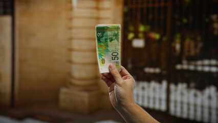 Hand holding israeli banknote outdoors in an urban setting showcasing modern business and...