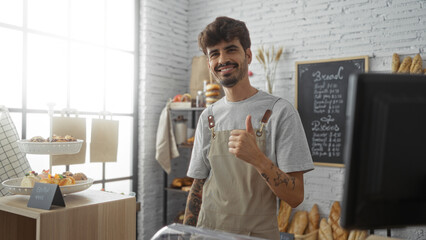 Handsome young man with beard smiling in a bakery shop interior wearing an apron and giving a thumbs up gesture