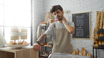Young man with beard and tattoos in a bakery rubbing his neck in discomfort, surrounded by bread and pastries, standing indoors by the counter.