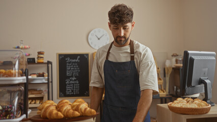 Young man working in a bakery shop surrounded by croissants, pastries, and a counter, with a clock and menu board in the background.