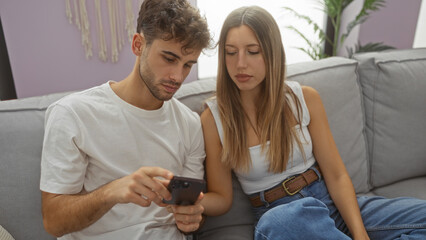 Couple sitting on sofa in cozy living room, man and woman looking at smartphone together, creating a warm and intimate home atmosphere.