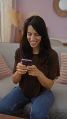Woman smiling at her smartphone while sitting in a cozy living room, capturing a joyful indoor moment in her home