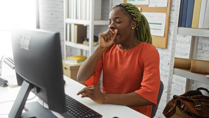 Woman yawning at computer in office setting represents fatigue during work hours with focus on young african american professional in casual attire and braided hairstyle.