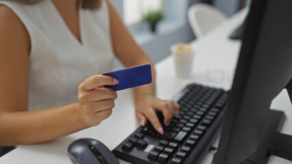 Woman using credit card in office for online payment, showing only hands, partial view, indoors, typing on keyboard, workplace setting with computer and coffee