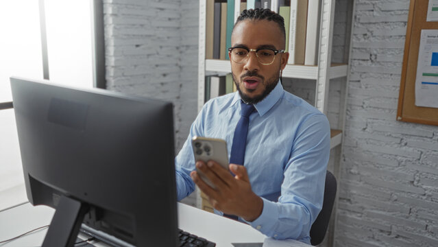 Young man with braids making a video call in a modern office environment, using a smartphone while wearing a formal blue shirt, surrounded by books and a computer. - Powered by Adobe